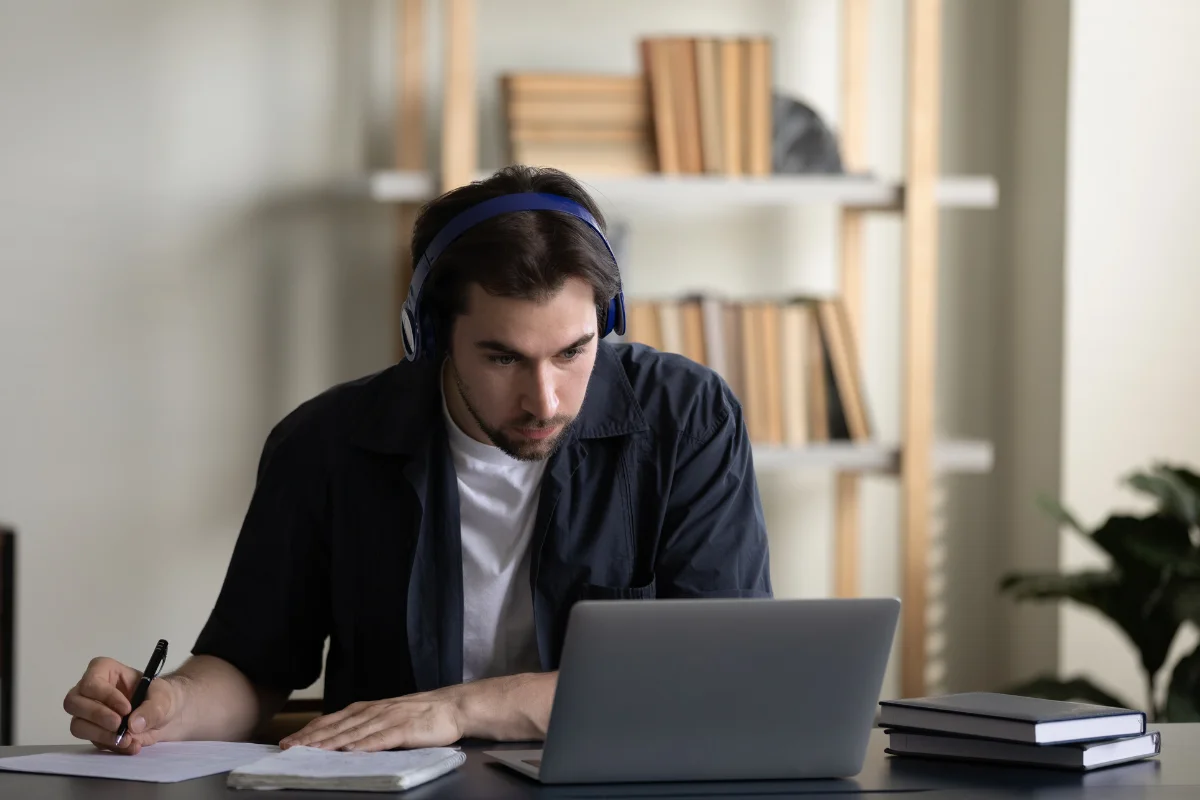 Homem concentrado, usando fones de ouvido, estudando no computador em uma mesa com anotações.