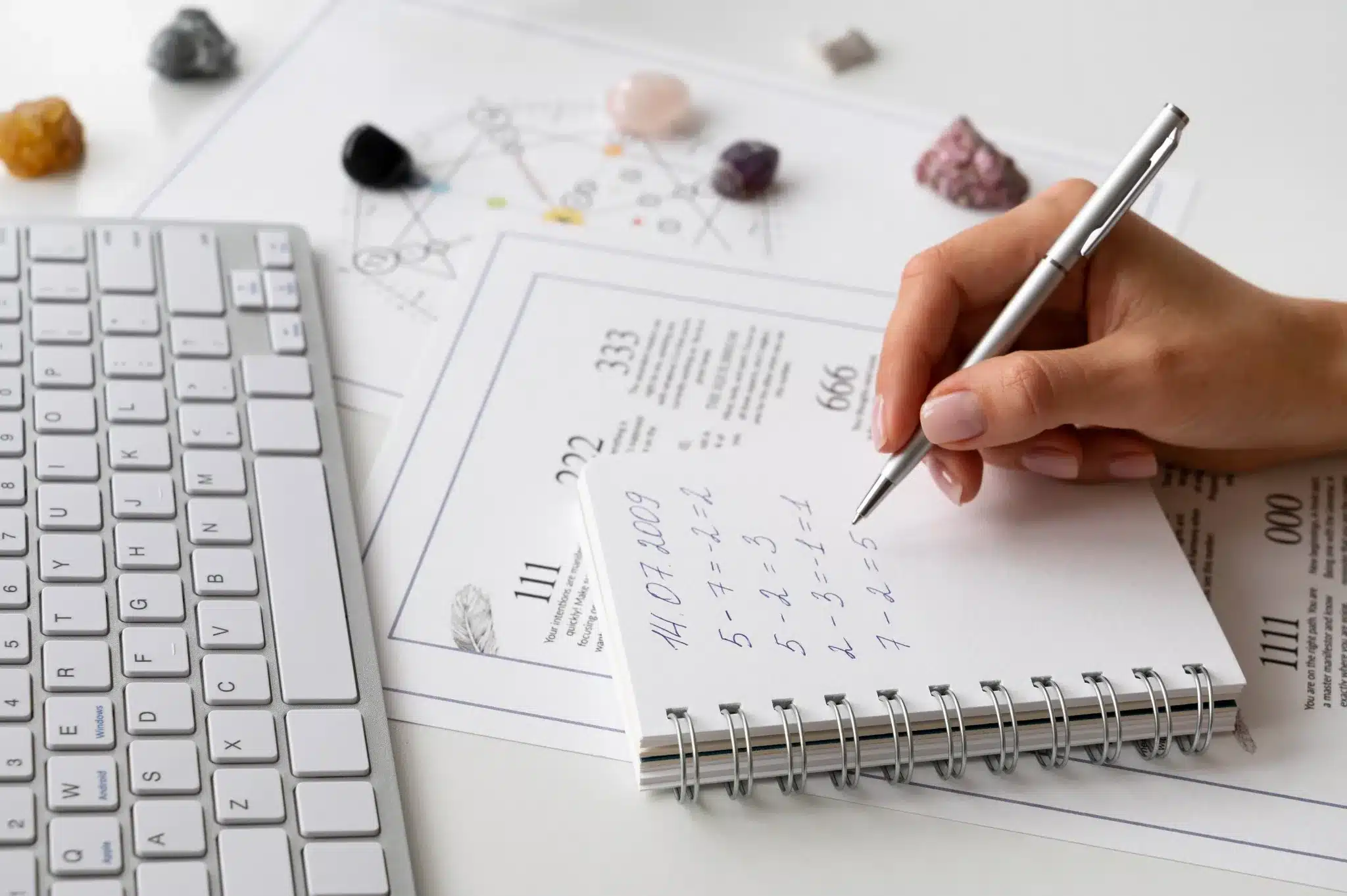 Mão escrevendo anotações sobre numerologia em um caderno ao lado de cristais e um teclado.