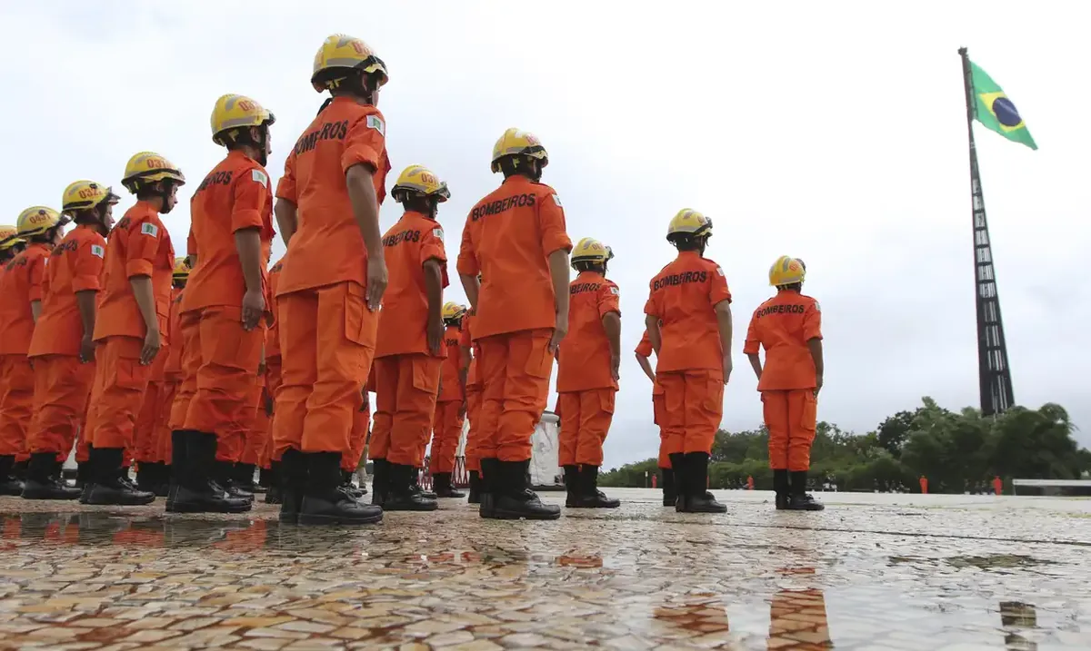 Bombeiros em formação durante cerimônia com bandeira do Brasil ao fundo.