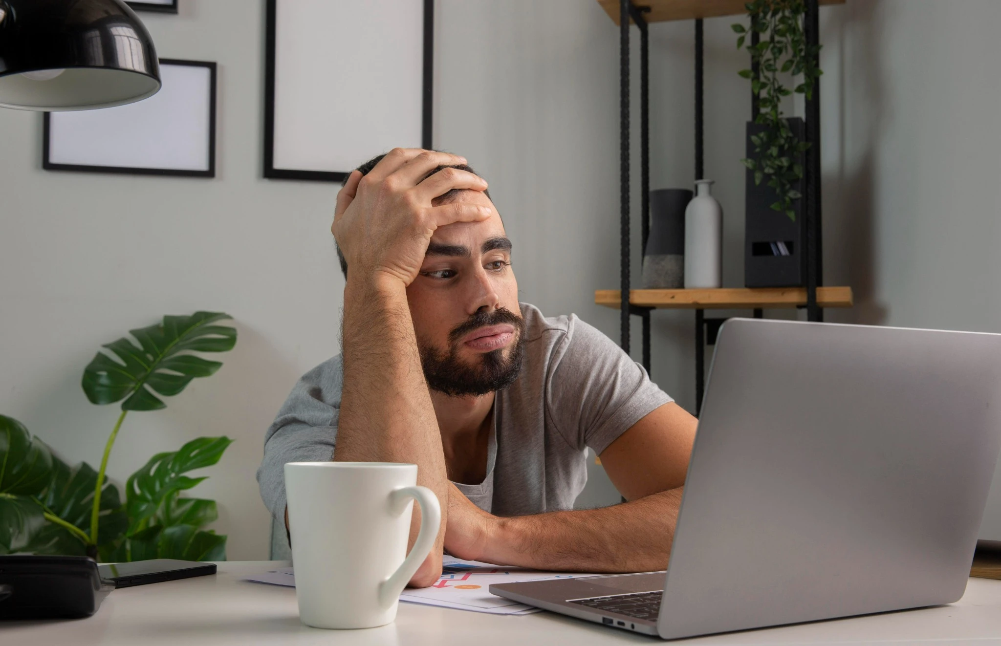 Homem com expressão cansada olhando para a tela do laptop enquanto apoia a cabeça na mão.