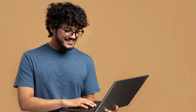 Homem sorridente usando óculos e camiseta azul, segurando e digitando em um notebook.