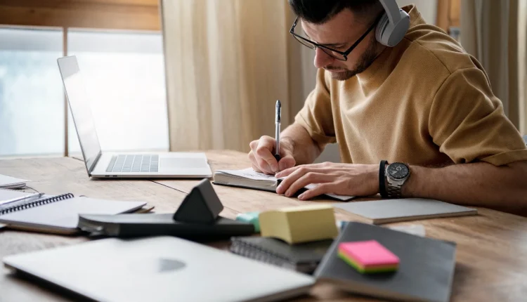Homem jovem usando fones de ouvido e óculos, escrevendo em um caderno durante estudo.