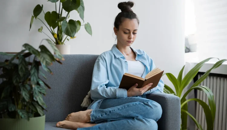Mulher concentrada lendo um livro em um ambiente confortável e iluminado, cercada por plantas verdes
