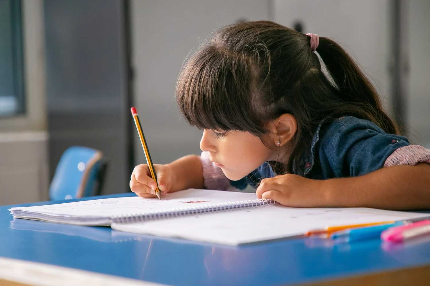 Menina concentrada escrevendo em caderno espiral, sentada à mesa azul em sala de aula.