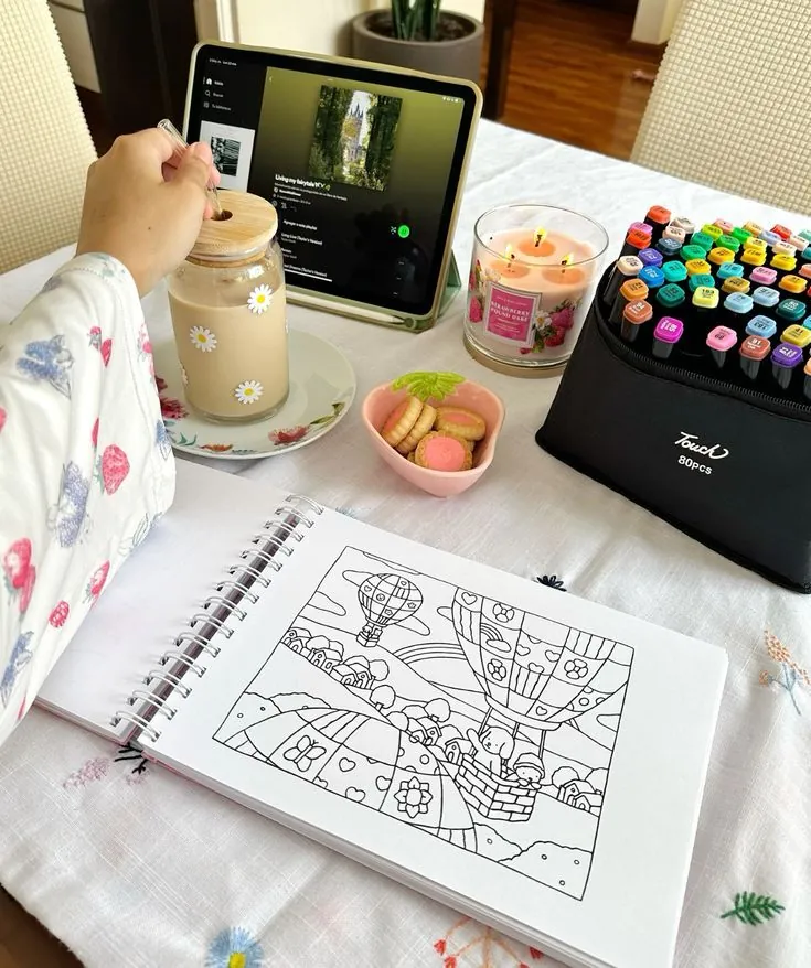 Mesa organizada com café, biscoitos, vela aromática, tablet com música e livro de colorir.