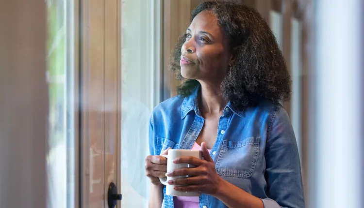 Mulher pensativa segurando uma caneca branca enquanto observa pela janela.