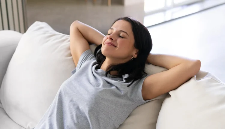 Mulher relaxando confortavelmente em um sofá branco, com expressão serena e os braços atrás da cabeça, lembrando de algo do passado.