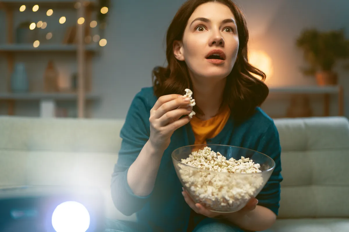 Mulher assistindo filme com expressão surpresa enquanto come pipoca.