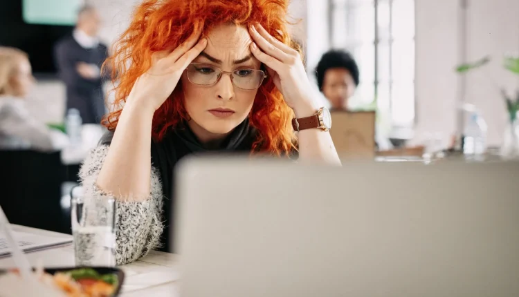 Mulher com cabelo ruivo, parecendo estressada, segurando a cabeça enquanto olha para o computador, em um escritório com pessoas ao fundo.