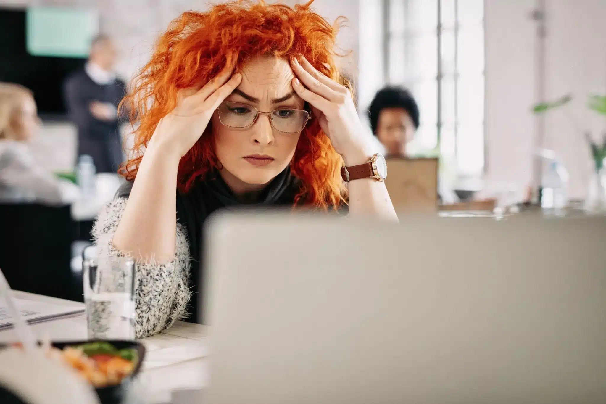 Mulher com cabelo ruivo, parecendo estressada, segurando a cabeça enquanto olha para o computador, em um escritório com pessoas ao fundo.
