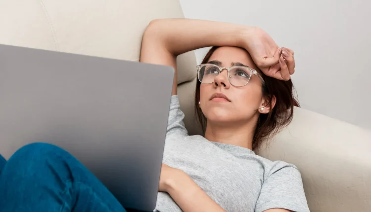 Mulher pensativa de óculos, deitada em um sofá segurando um laptop.