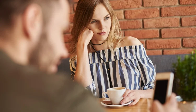 Mulher pensativa e séria segurando uma xícara de café enquanto homem ao fundo está distraído com o celular.