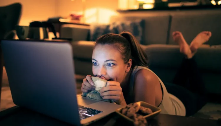 Mulher jovem deitada no chão, assistindo atentamente ao laptop com expressão animada, segurando uma manta.