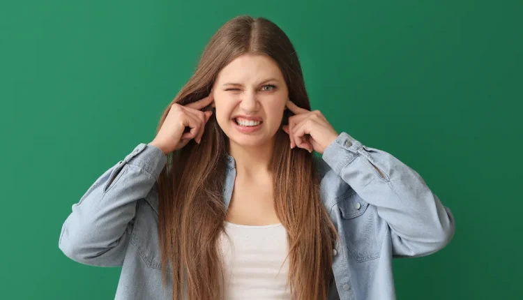 Mulher jovem tampando os ouvidos com os dedos e expressão de incômodo sobre fundo verde.