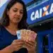 Mulher confere cédulas de reais em frente a uma agência da Caixa, representando o início dos pagamentos do Bolsa Família de julho.