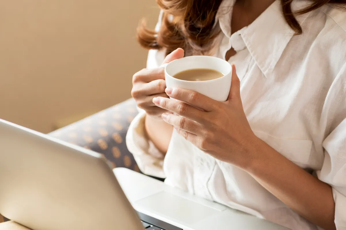 Mulher segurando uma xícara de café com leite enquanto trabalha no notebook.