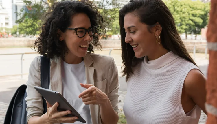 Duas mulheres sorrindo enquanto visualizam conteúdo em um tablet, refletindo a importância de hábitos para o sucesso profissional e financeiro.
