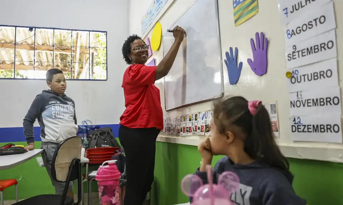 Professora escreve no quadro branco enquanto crianças observam em sala de aula colorida.