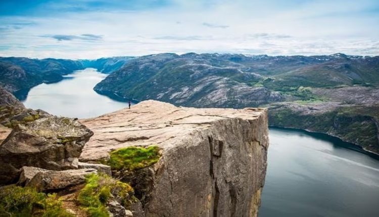 Teste de QI com pessoa em pé na beira de um penhasco com vista para um fiorde rodeado por montanhas e água.