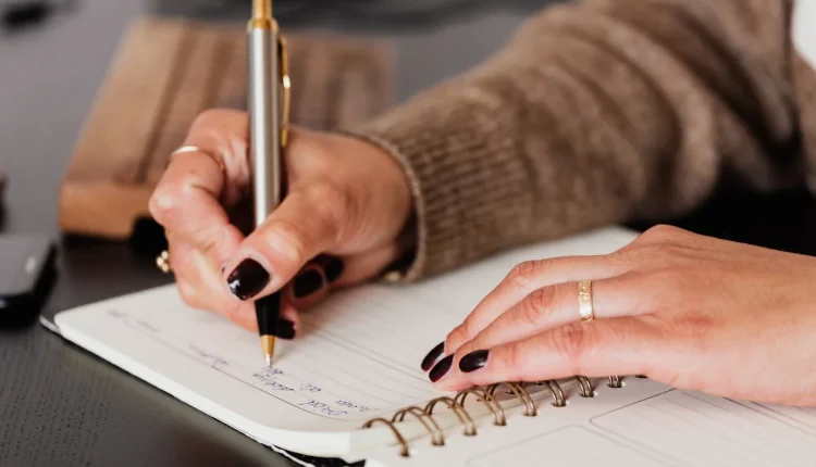 Pessoa escrevendo em caderno, utilizando uma caneta, com uma mesa ao fundo.