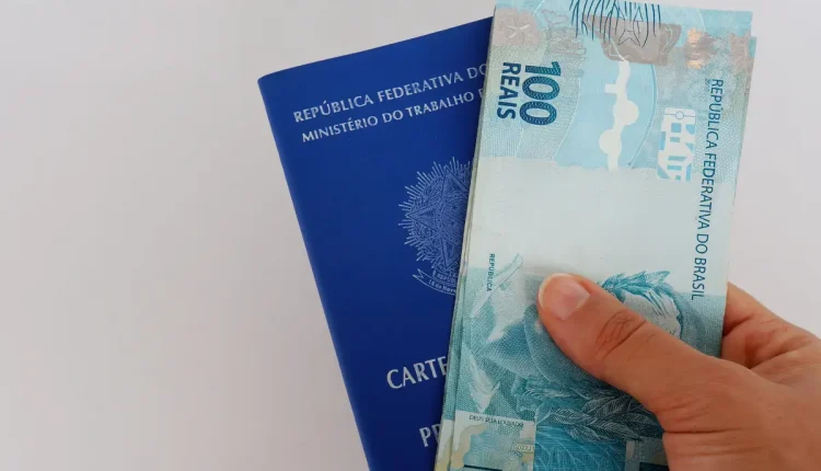 Mão segurando cédulas de 100 reais e carteira de trabalho brasileira.