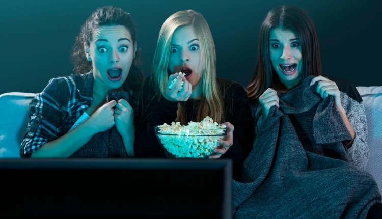 Três amigas assistindo a um filme de terror, com expressões de medo, segurando pipoca e cobertores.