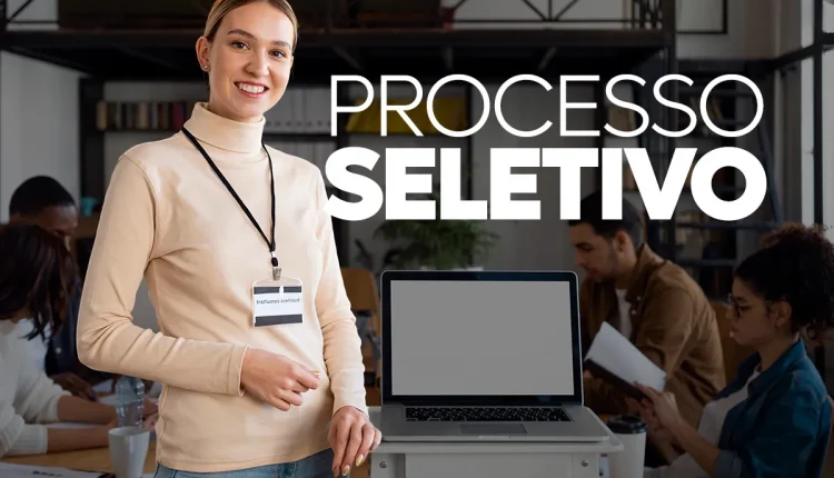Imagem de uma mulher sorridente usando crachá, com laptop e pessoas ao fundo em um ambiente de trabalho, representando o conceito de processo seletivo.