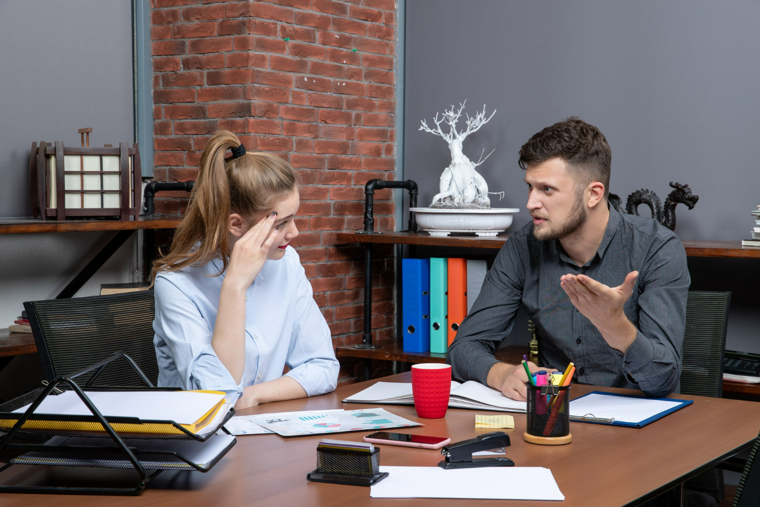 Homem falando de forma intensa para colega de trabalho que parece preocupada, em ambiente de escritório moderno.