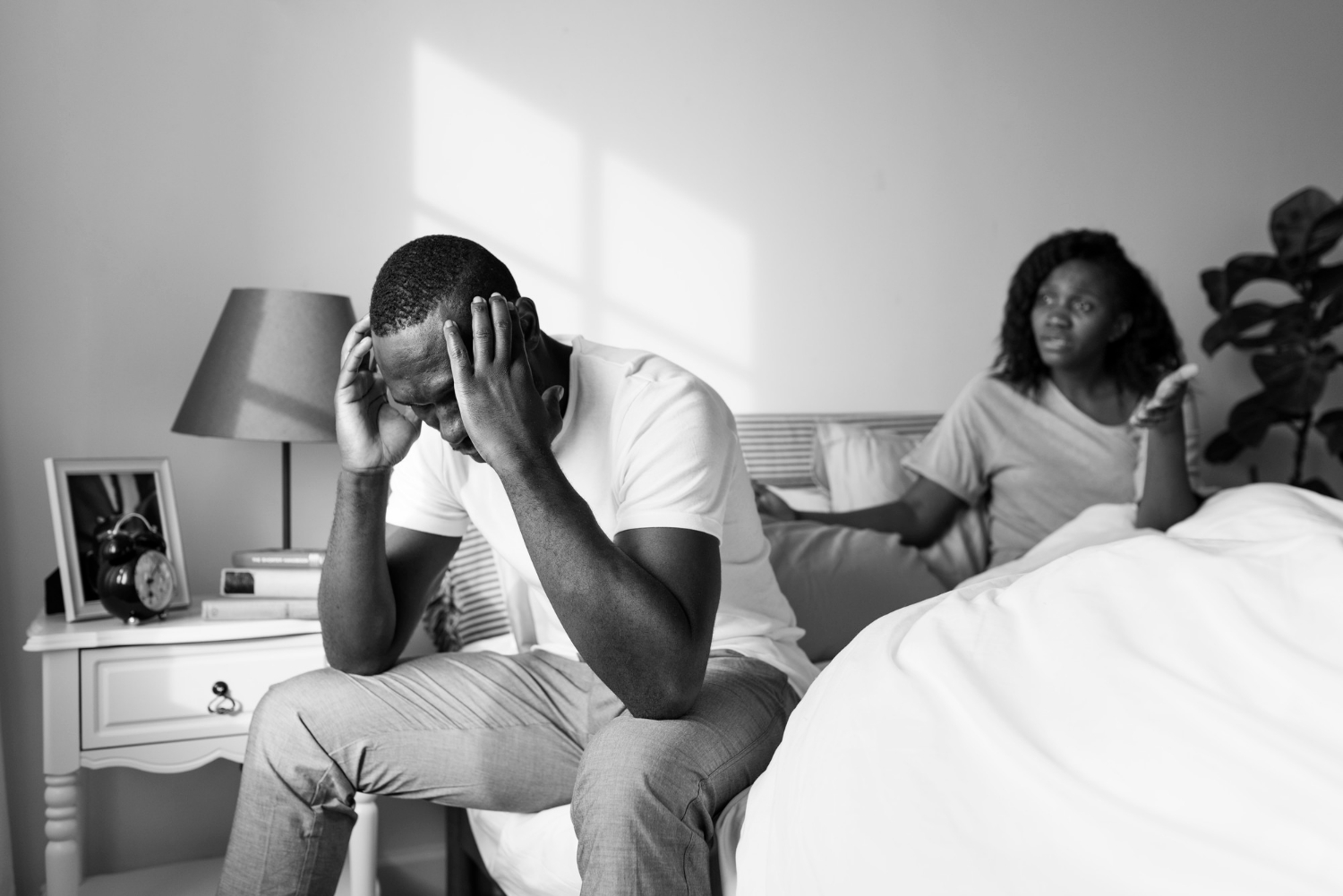 Homem com expressão de angústia sentado na beira da cama enquanto mulher fala ao fundo, simbolizando conflitos e desafios em relacionamentos devido a problemas pessoais.