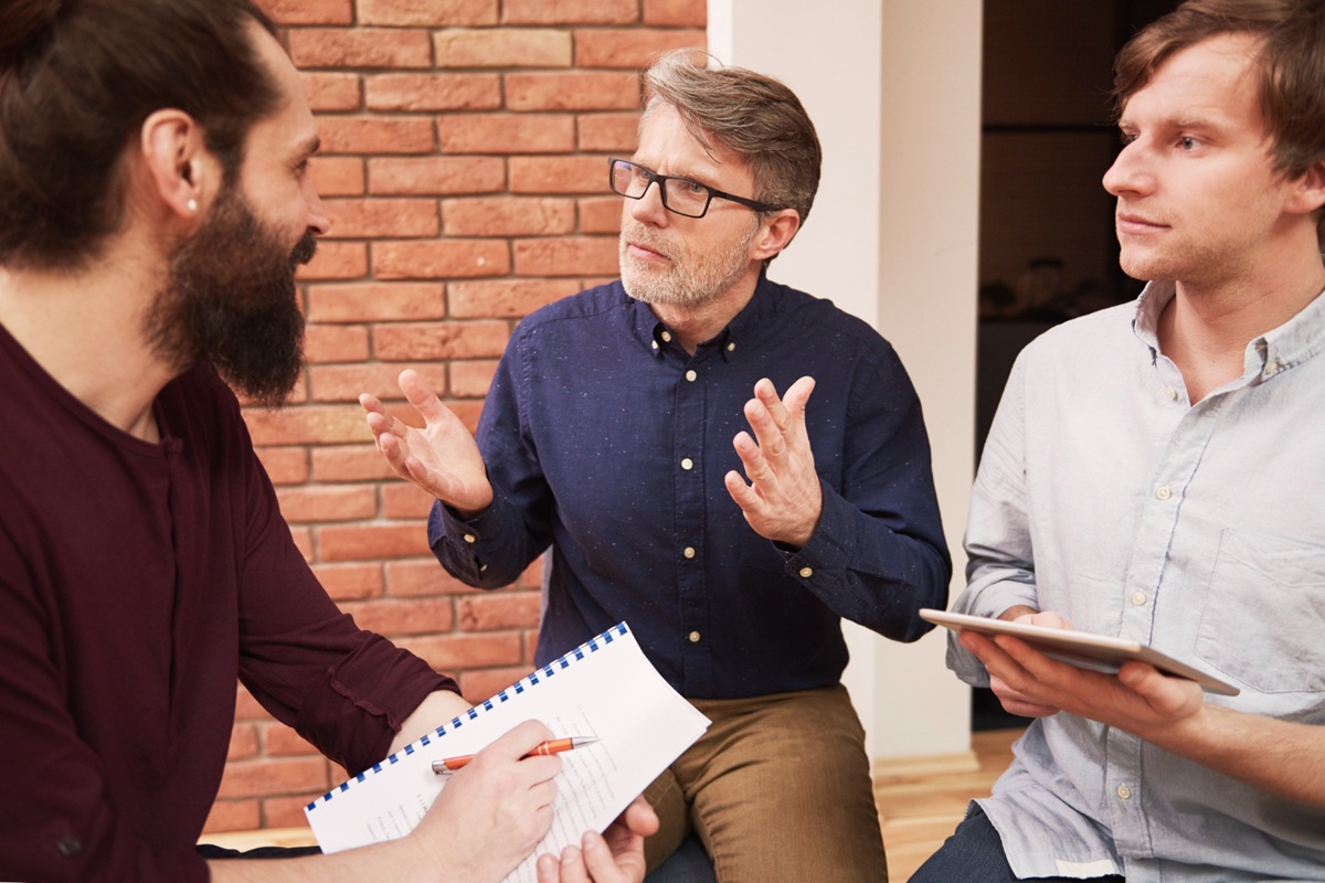 Três homens discutindo ideias com bloco de anotações em ambiente interno.