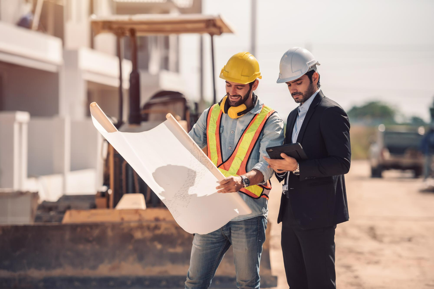 Dois engenheiros em canteiro de obras, um com capacete e colete refletivo, examinando um plano de construção enquanto o outro usa tablet.