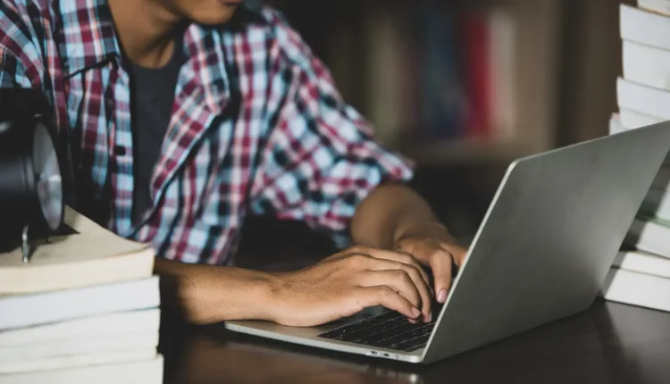 Jovem em um ambiente de estudo, digitando no laptop enquanto livros estão ao seu redor, simbolizando a dedicação aos estudos para cursos técnicos.