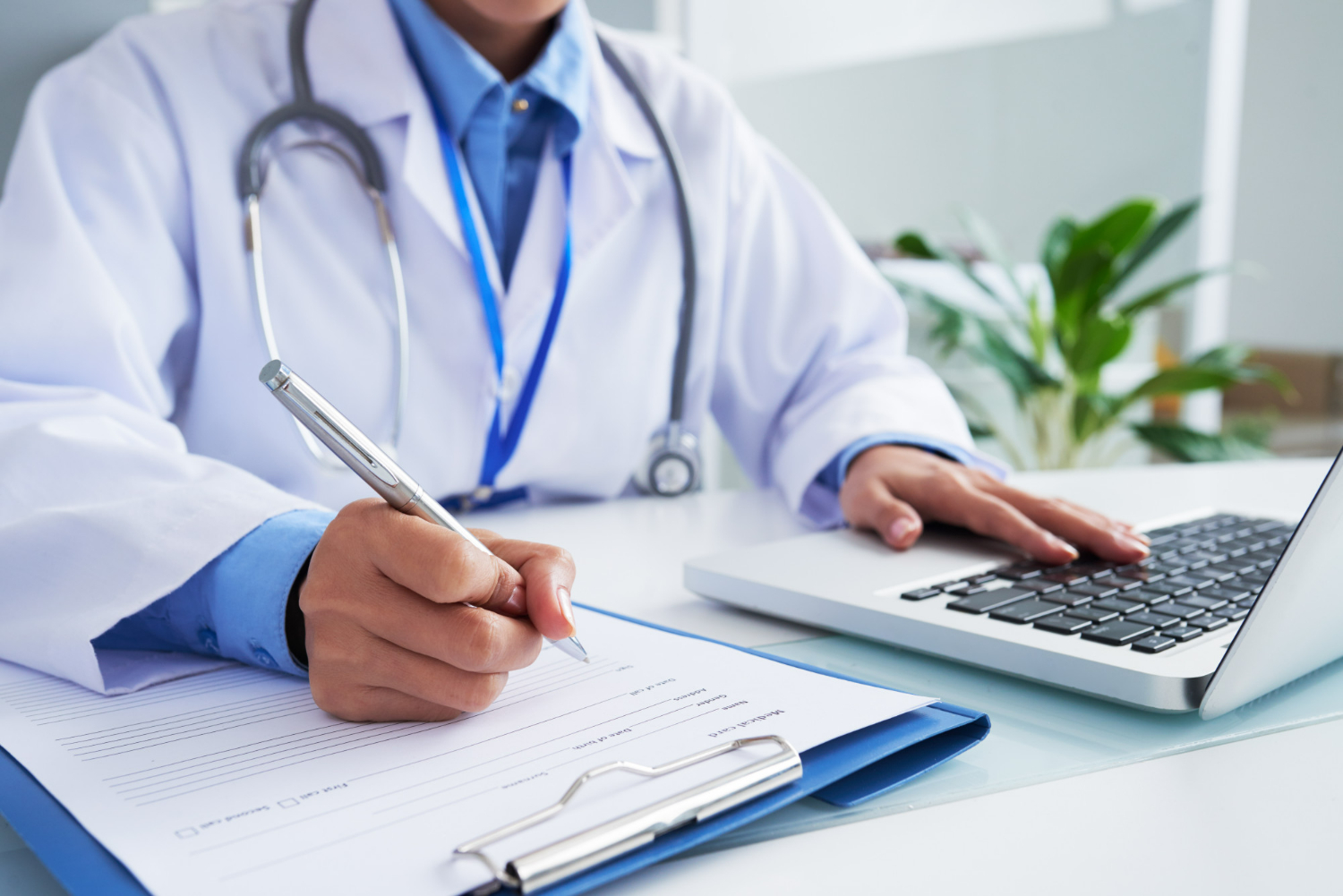 Médico com jaleco e estetoscópio anotando informações em prontuário ao lado de computador.