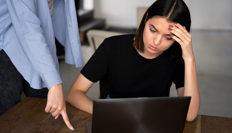 Jovem mulher preocupada em frente ao laptop enquanto outra pessoa aponta com o dedo, simbolizando pressão ou cobrança no trabalho.