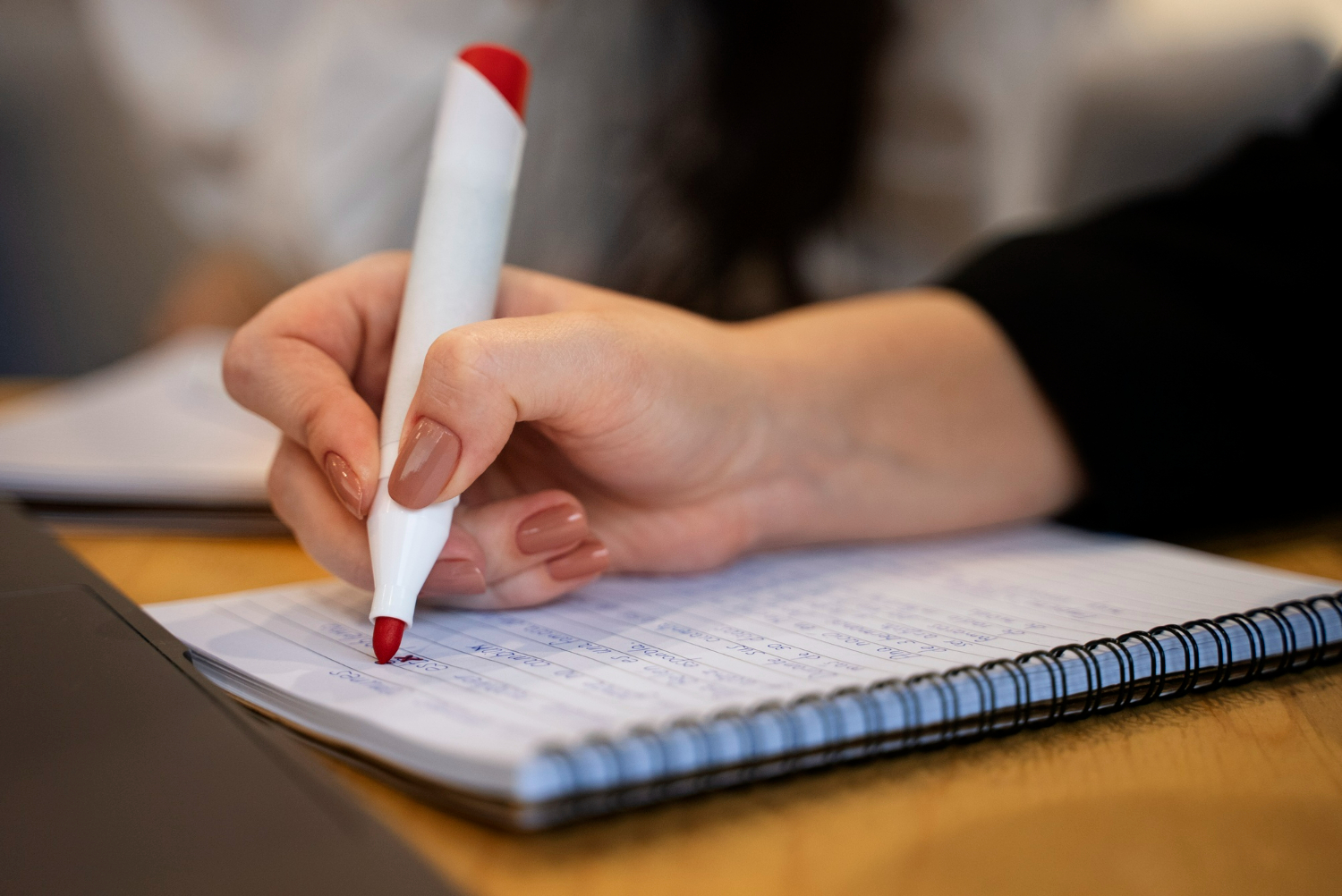 Pessoa escrevendo com caneta vermelha sobre caderno espiral, representando o ato de corrigir e revisar a grafia correta.