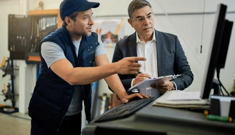 Dois profissionais conversando em um ambiente industrial, com um operador explicando algo para o supervisor em frente a um computador.