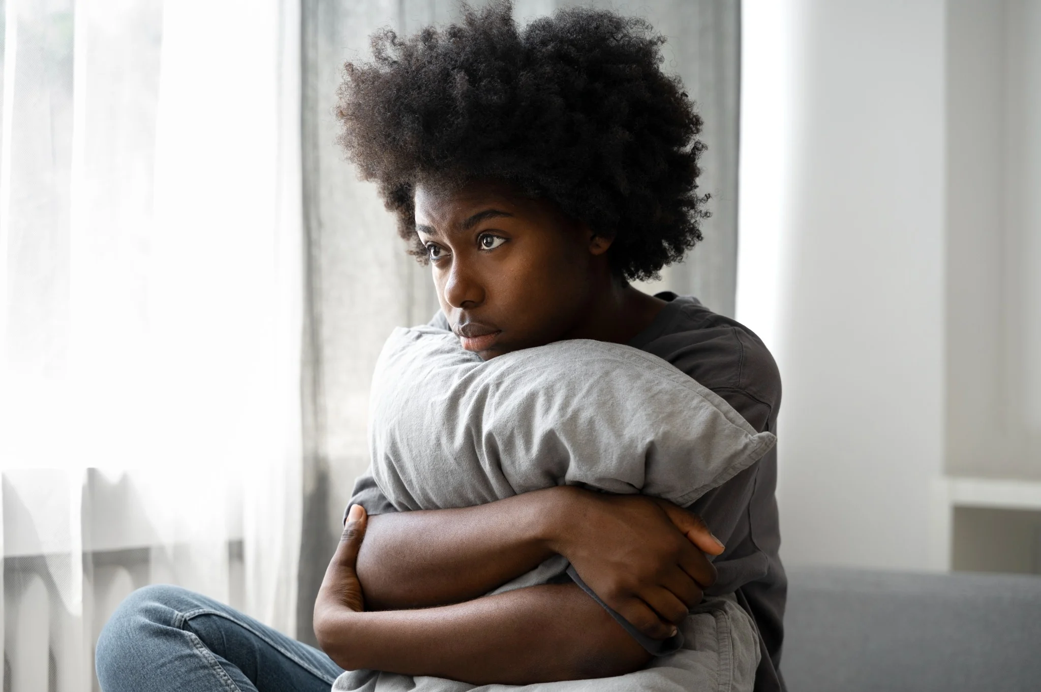Jovem com cabelo afro segurando um travesseiro, com expressão pensativa e triste, sentado em um sofá em um ambiente doméstico.