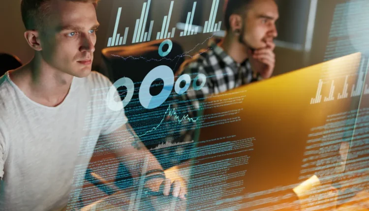 Dois profissionais analisando dados em tempo real com gráficos e códigos na tela.