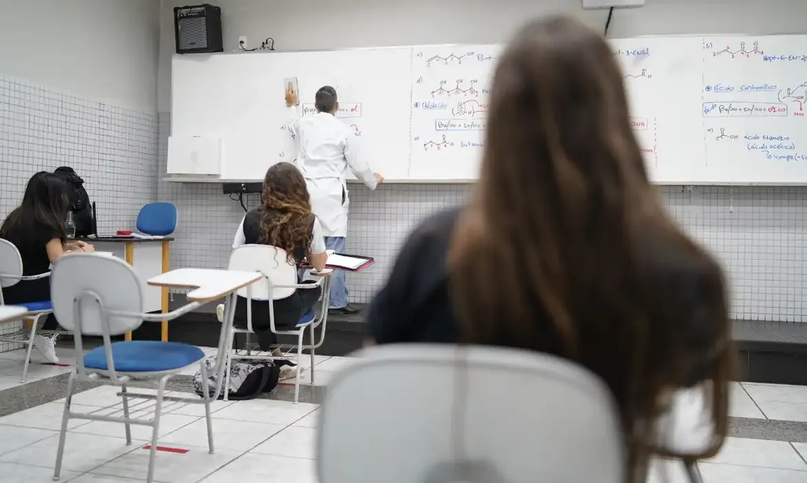 Alunos assistem aula em escola pública com professor escrevendo na lousa.