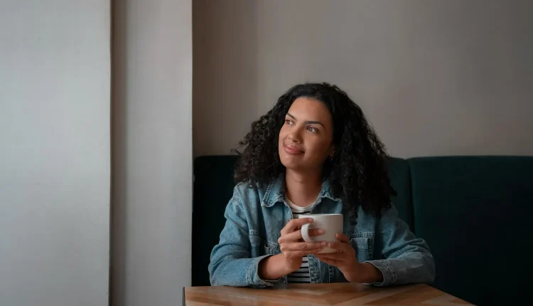 Mulher sorrindo enquanto segura uma xícara de café, refletindo sobre seus pensamentos.