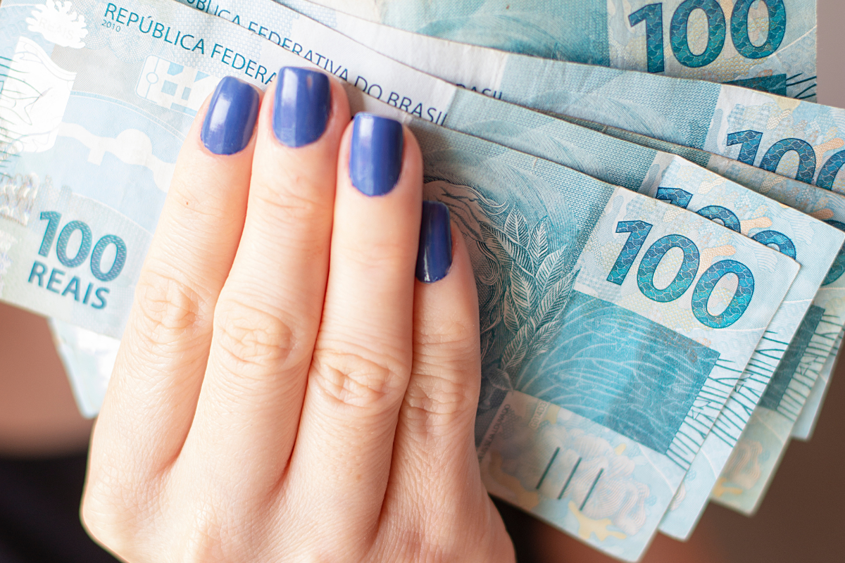 Mão feminina segurando cédulas de 100 reais com unhas pintadas de azul.