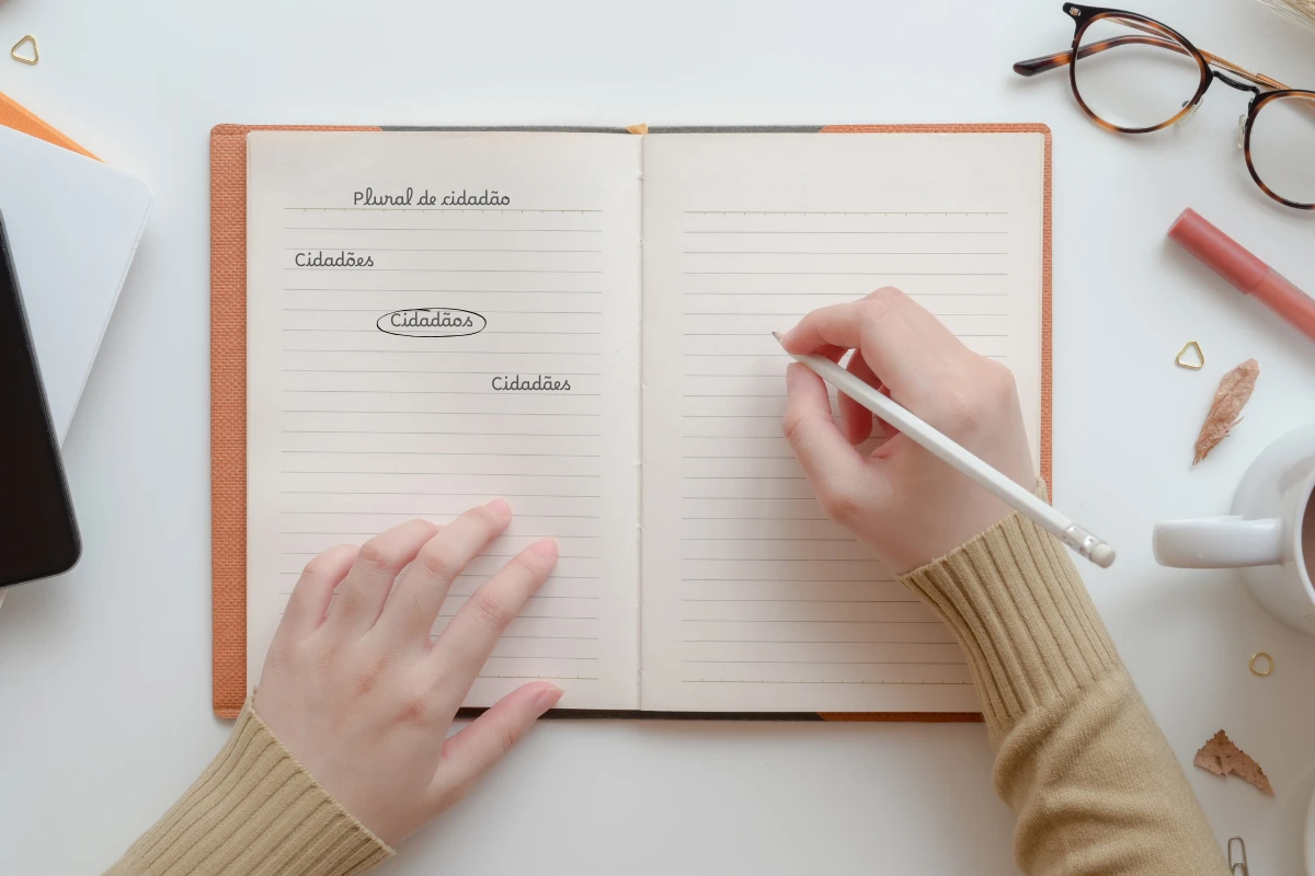 Mãos femininas escrevendo em um caderno aberto com anotação sobre o plural da palavra cidadão.