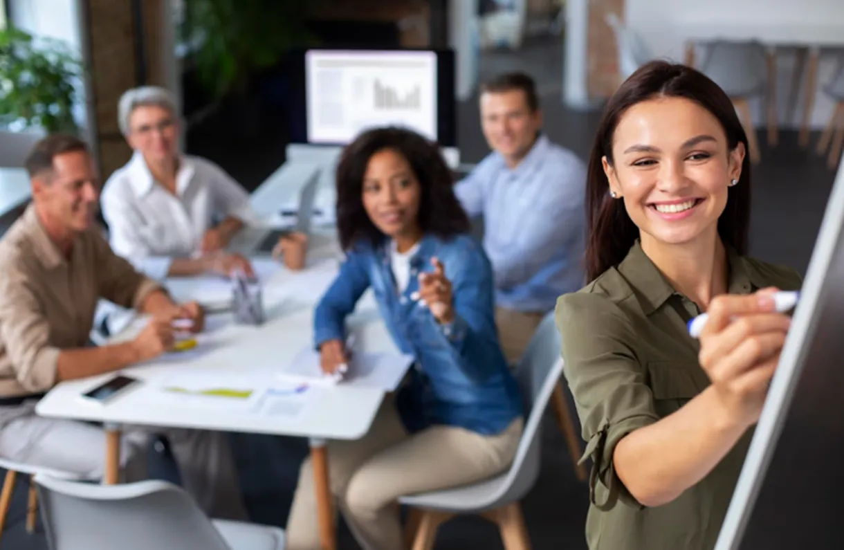 Profissionais colaborando em uma reunião, com uma mulher apresentando uma ideia na lousa, simbolizando habilidades de comunicação em equipe.