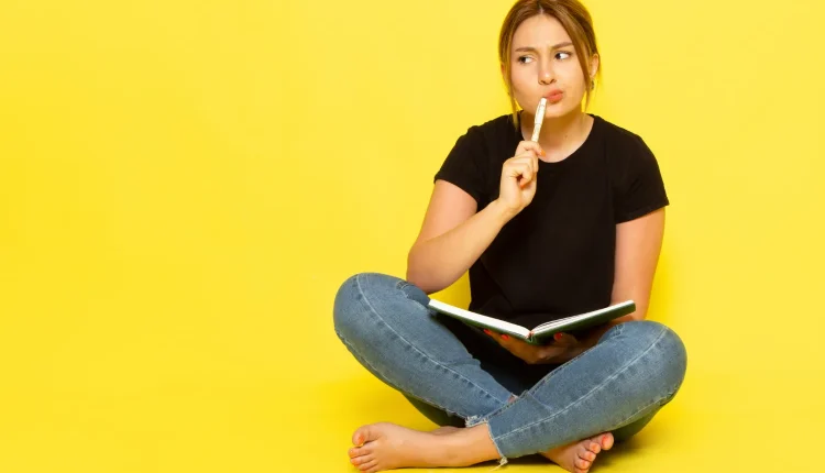 Mulher sentada com um livro na mão, pensativa e com um gesto de dúvida, ilustrando a confusão sobre 'Deus te abençoe' ou 'Deus te abençõe'.