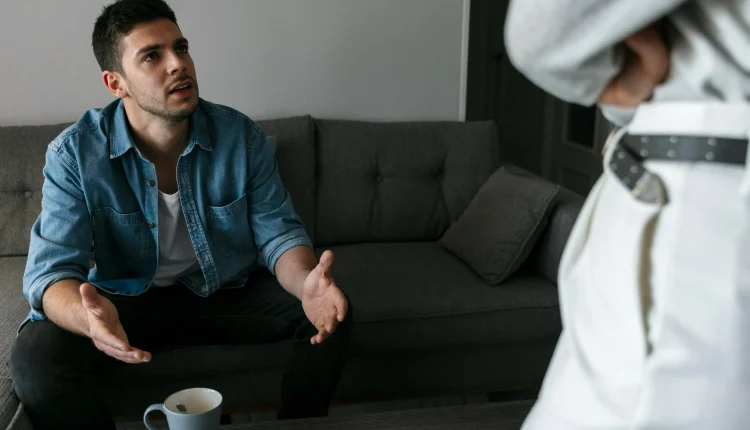 Homem sentado no sofá, conversando de forma séria com outra pessoa em pé, expressão de explicação ou argumentação.