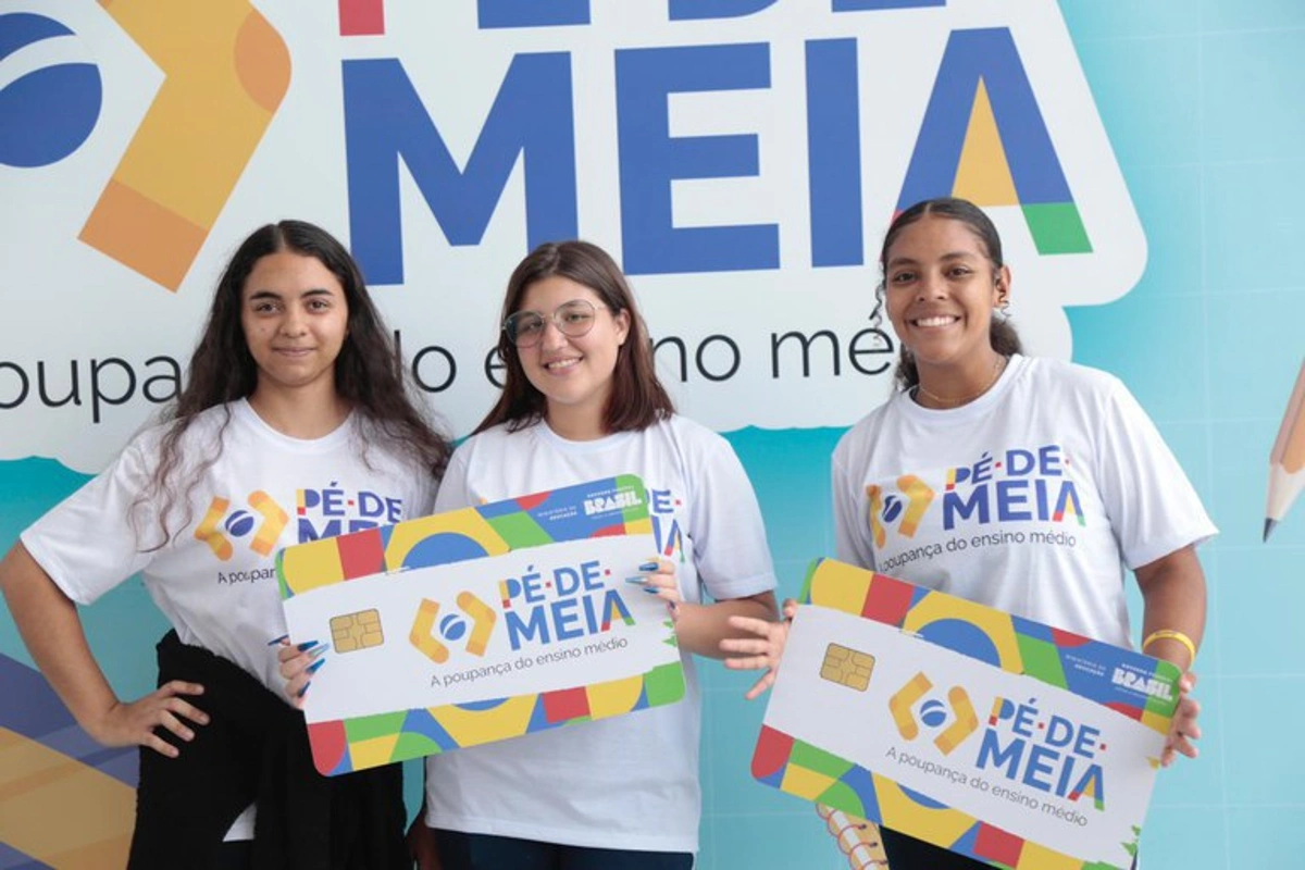 Três estudantes sorrindo segurando cartões grandes do programa Pé de Meia, vestindo camisetas do projeto.