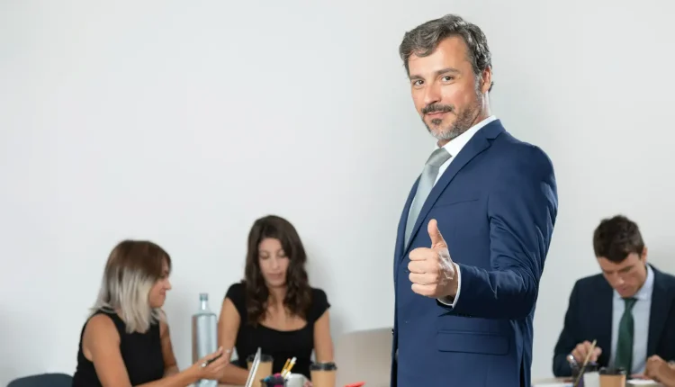 Homem de terno azul com expressão confiante, interagindo com sua equipe em ambiente de escritório moderno, promovendo comunicação e liderança.