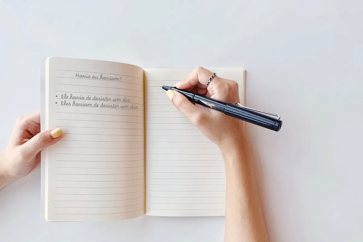 Pessoa escrevendo em um caderno com caneta preta, focando na diferença entre 'Havia' e 'Haviam'.