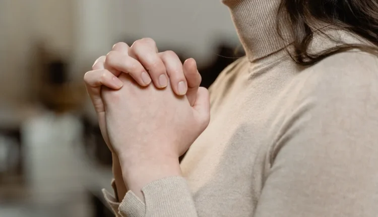 Mãos de uma pessoa entrelaçadas em oração, com fundo desfocado em ambiente interno.