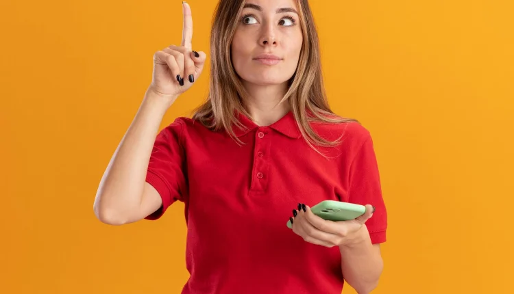 Mulher jovem com camisa vermelha segurando celular e apontando o dedo indicador para cima, simbolizando uma ideia ou atenção.